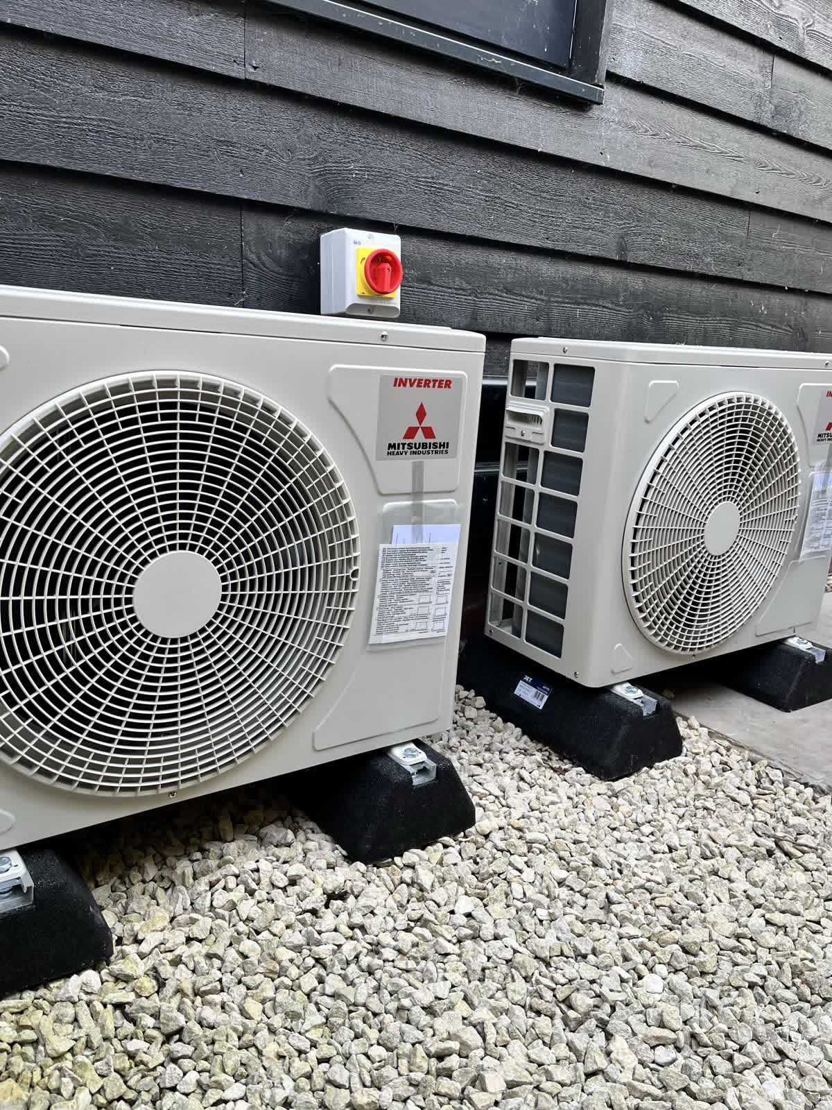 Two outdoor air conditioning units mounted on anti-vibration feet against dark timber cladding